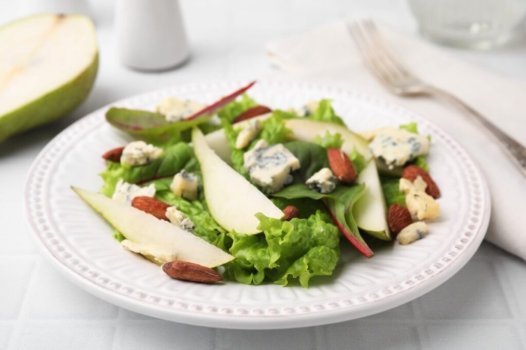 Caramelized Pear, Blue Cheese, and Honey-Roasted Almond Salad: Salad with pear slices, blue cheese, lettuce, almonds on a white plate with a fork and napkin nearby.