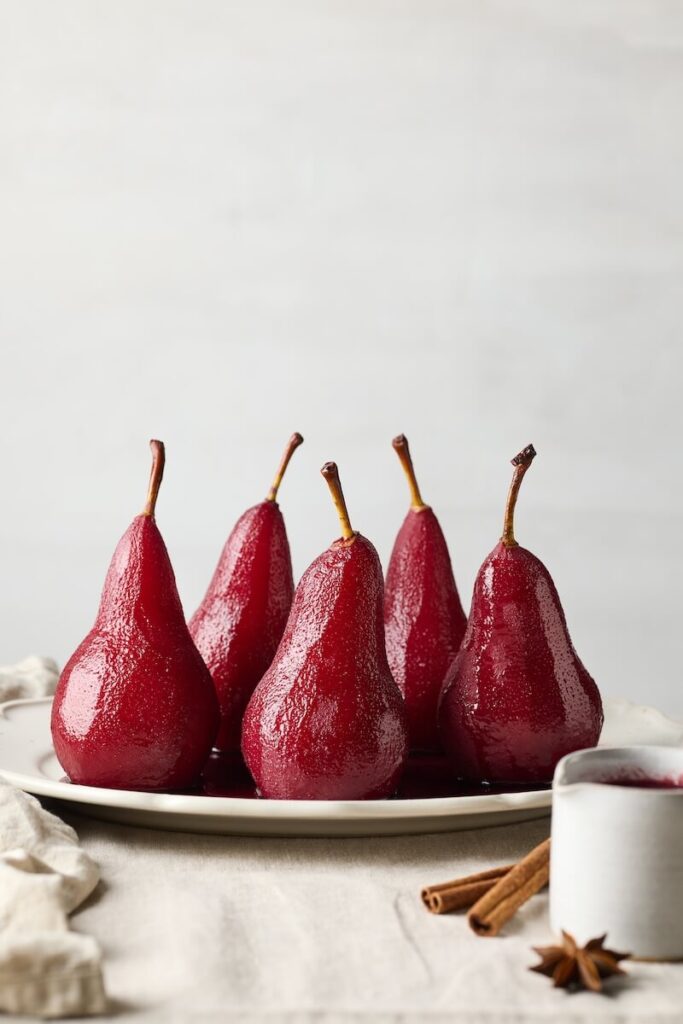Basic Poached Pears: Six red poached pears on a white plate, with cinnamon sticks and star anise nearby on a beige tablecloth.
