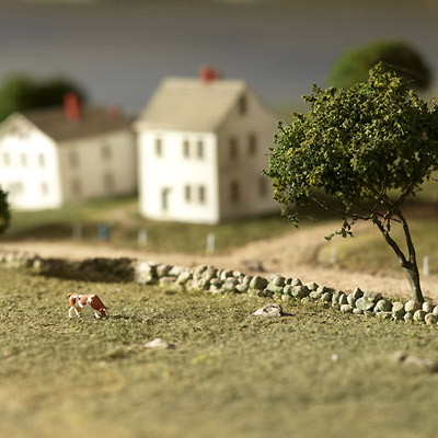 Model of a rural landscape featuring small houses, a tree, a stone wall, and a grazing cow in the foreground.