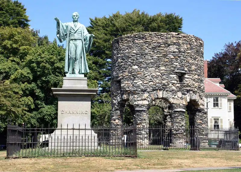 A green statue labeled "Channing" stands beside an old stone tower with arches, both surrounded by a black metal fence on a grassy area with trees and a house in the background.