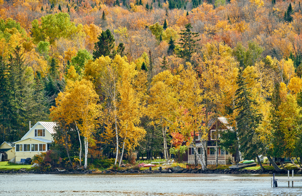 The 25 Best Foliage Towns in New England. Two houses by a lake surrounded by vibrant autumn trees with colorful foliage.