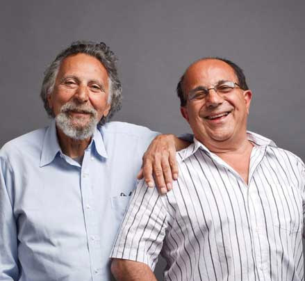 Two older men stand closely together, both smiling. One man has his arm around the other's shoulder. The background is plain gray.