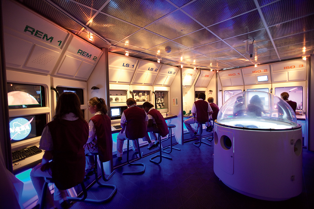 Children in uniforms sit at computer stations in a space-themed control room with NASA logos and blue lighting.