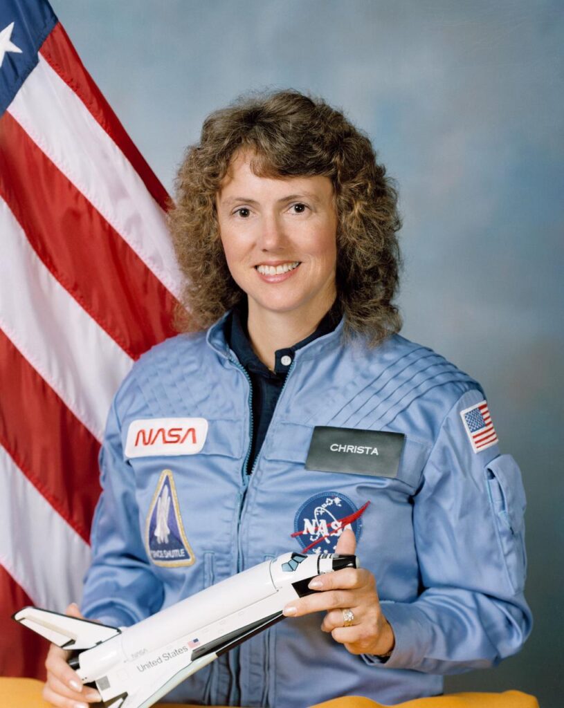 Woman in a blue NASA jumpsuit holding a space shuttle model, with an American flag behind her.