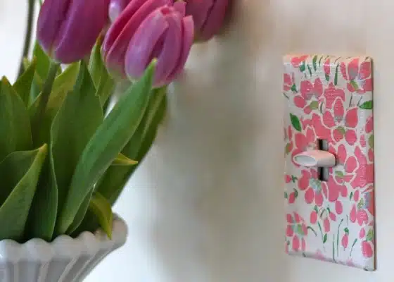 Close-up of a floral-patterned light switch plate on a wall, with a bouquet of purple tulips in a white vase nearby.
