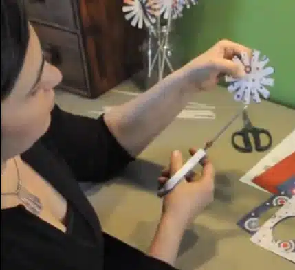 A person holding and trimming a paper snowflake with scissors, sitting at a table with more paper snowflakes, scissors, and other craft supplies.