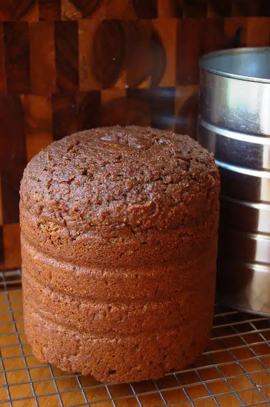 A cylindrical loaf of brown bread with a textured surface is cooling on a wire rack next to a silver tin. The background is a wooden surface with a checkerboard pattern.