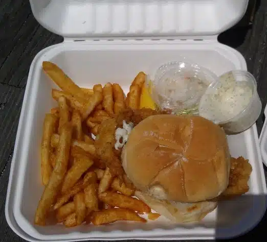A takeout container with a fried chicken sandwich, seasoned fries, a side of coleslaw, and a container of dipping sauce.