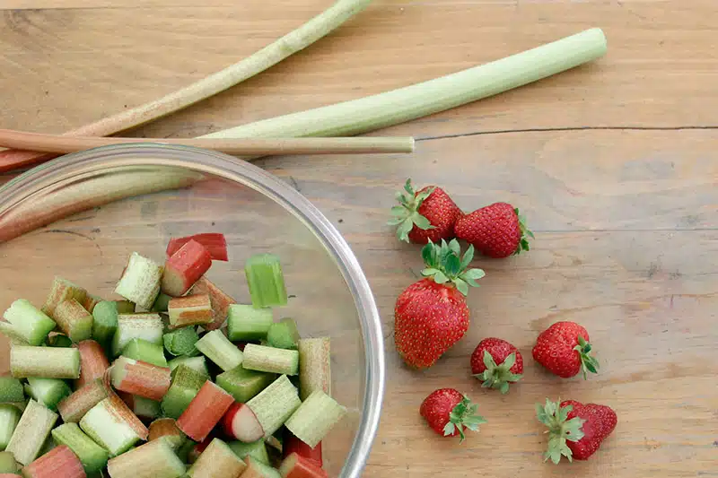 Chopped rhubarb in a glass bowl next to fresh rhubarb stalks and strawberries on a wooden surface.