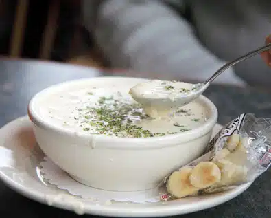 A bowl of creamy soup garnished with herbs is placed on a plate. A spoon is being used to scoop the soup. A packet of oyster crackers is next to the bowl.