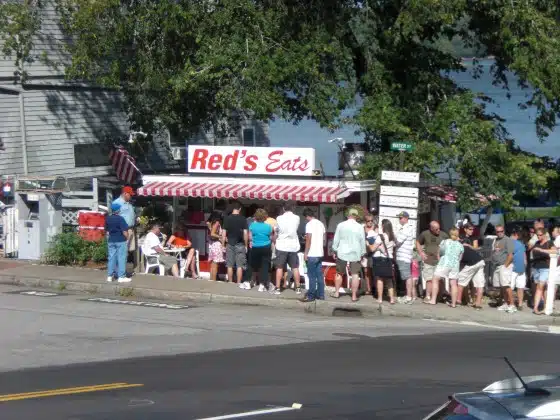 A group of people wait in line outside Red's Eats, a small eatery with a striped awning, located near a road and partially surrounded by trees.