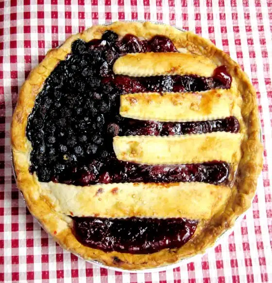 A berry pie with a lattice crust design resembling the American flag, placed on a red and white checkered tablecloth.