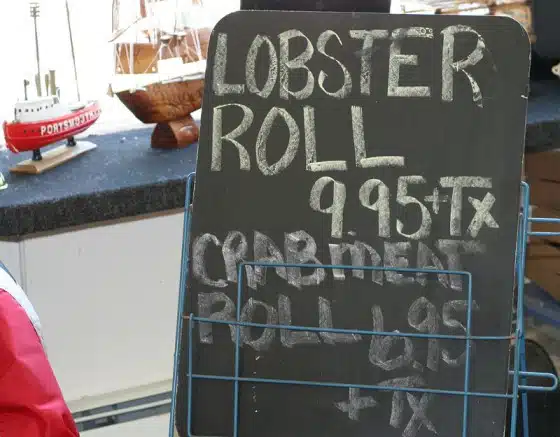 A chalkboard sign advertises "Lobster Roll 9.95 + Tx" and "Crabmeat Roll 8.95 + Tx" in front of model ships on a counter.