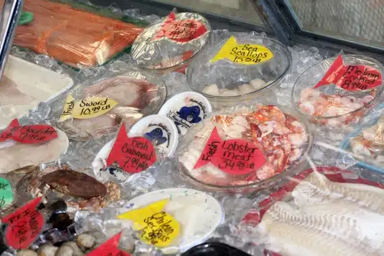 A seafood display with labels showing prices for various items, including lobster meat, sea scallops, swordfish, and salmon, among others.