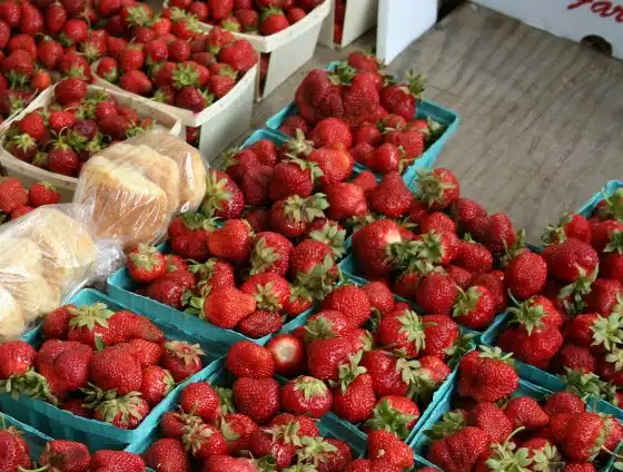 Punnets of fresh strawberries are displayed for sale at a market, with some packaged rolls visible to the left.