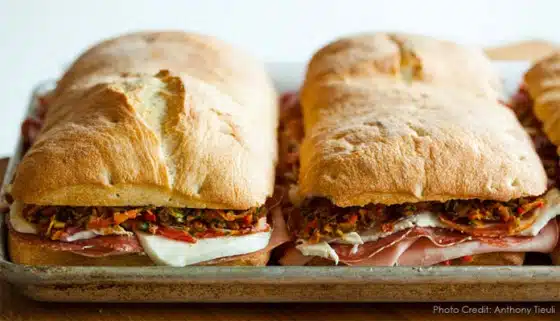 Two large sandwiches filled with meats, cheese, and chopped vegetables are placed side by side on a metal tray.