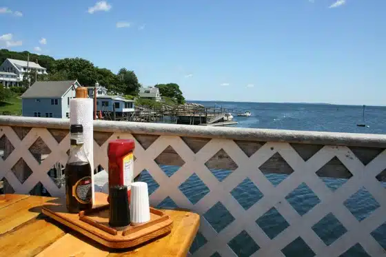 A wooden table with condiments and napkins next to a lattice railing overlooks a waterfront with houses, a dock, and a large body of water under a partly cloudy sky.