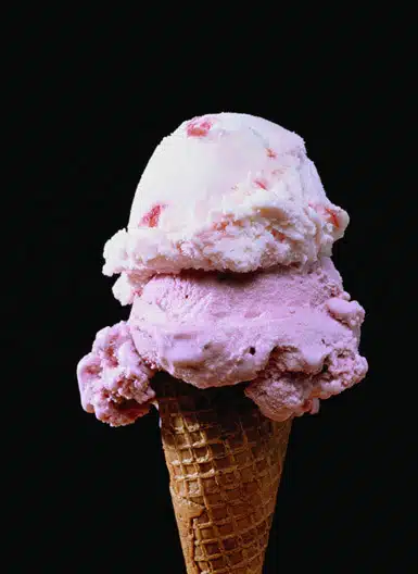 Two scoops of pink ice cream, one light and one darker, on a waffle cone against a black background.