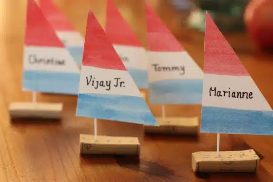 Small model sailboats with name tags on paper sails, placed on a wooden table. The names visible are "Vijay Jr.", "Tommy", and "Marianne.