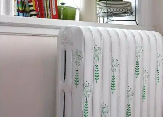 A white radiator decorated with green floral stencil patterns is positioned against a wall, with shelves of books, a teapot, and stacked plates visible in the background.