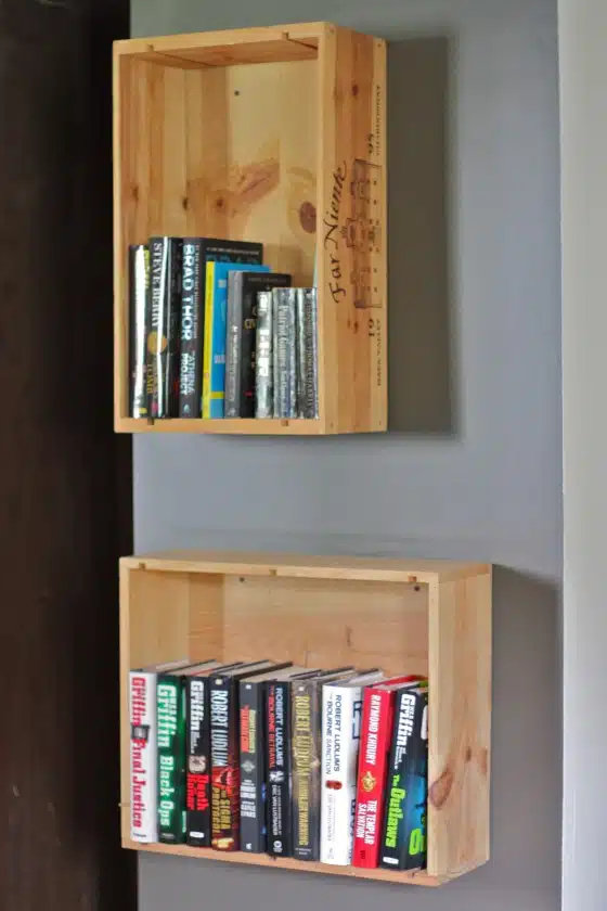 Two wooden crates mounted on a wall used as bookshelves, each holding a variety of books. The top shelf has a mix of genres, while the bottom shelf appears to contain mostly thriller novels.