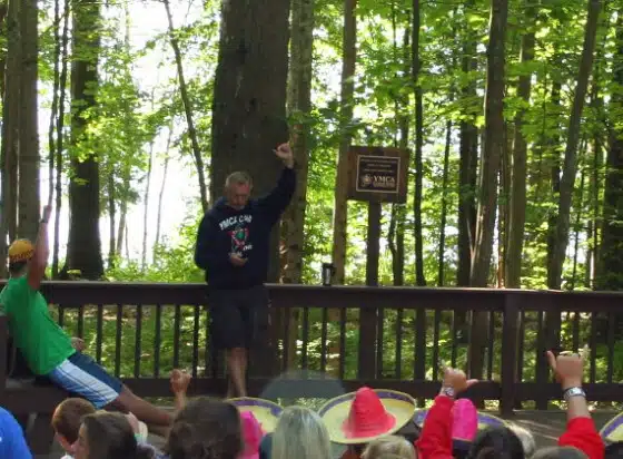 A person stands on a wooden platform raising their arm while speaking to a group of people in an outdoor, wooded area.