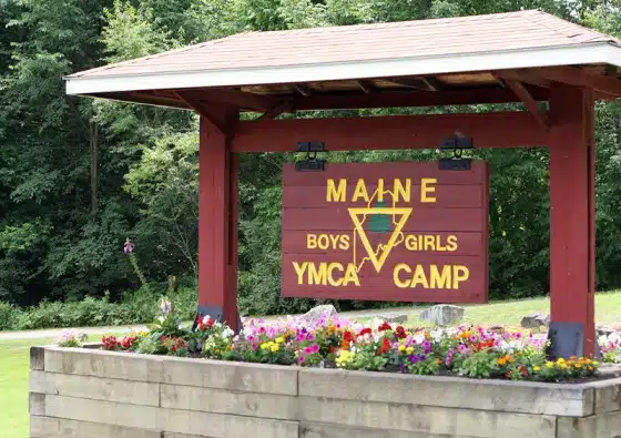 A wooden sign under a small roof reads "Maine YMCA Boys & Girls Camp." It is surrounded by a flower bed with colorful flowers. Trees are visible in the background.