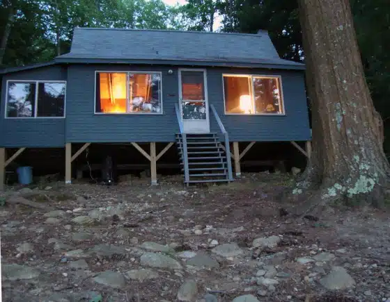 A blue wooden cabin with illuminated windows, elevated on stilts, surrounded by trees and rocky ground, viewed from the front.