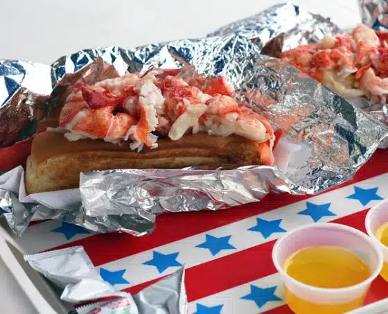 Two lobster rolls wrapped in foil, placed on a tray with a red, white, and blue star pattern. There are small cups of melted butter and condiment packets on the tray.