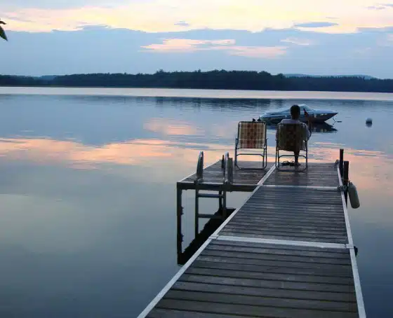 A person sits on a chair at the end of a wooden dock facing a calm lake with a small boat tied nearby, during sunset.