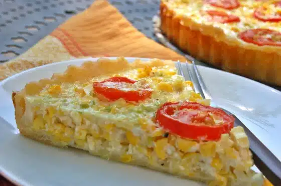 A slice of corn and tomato quiche on a white plate with a fork, with the rest of the quiche in the background.