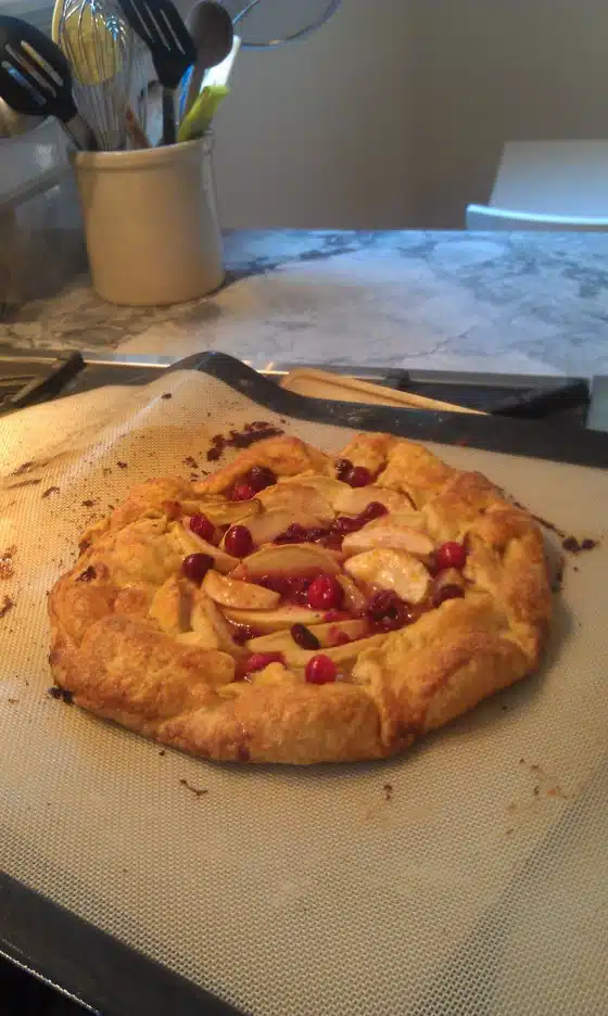 A freshly baked fruit galette with a golden-brown crust sits on a baking sheet atop a silicone mat in a kitchen. The galette contains slices of fruit and red berries.