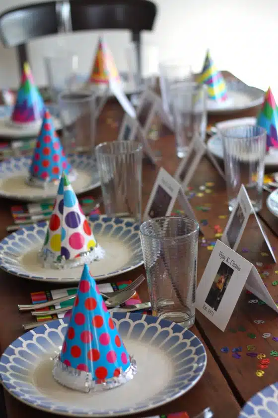 A table set for a party with colorful party hats on plates, clear glasses, and name cards. Confetti is scattered on the table.