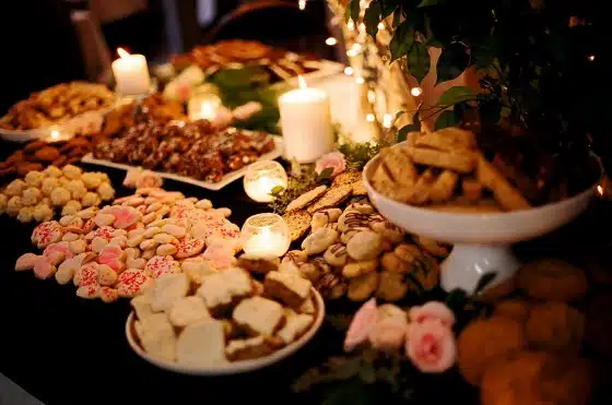 An assortment of various cookies and pastries displayed on several platters, with decorative candles and greenery in the background.