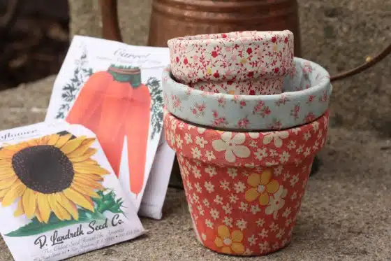 Three flower pots with floral fabric covers are stacked beside seed packets for carrots and sunflowers on a stone surface. An old watering can is in the background.