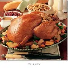 A roasted turkey on a platter surrounded by vegetables, apple slices, and garnishes. Background includes a bowl of stuffing, a bowl of cranberries, a loaf of bread, and various sauces.