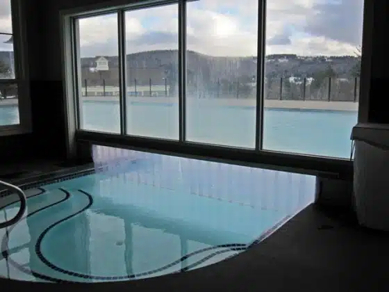 Image of an indoor pool area with large windows looking out onto an outdoor pool and scenic hills. The setting is clean and spacious with a calm atmosphere.