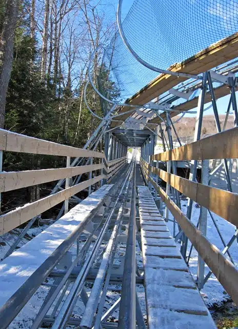 Steel track of an alpine coaster with wooden guardrails and a protective netting, extending through a forested area under clear blue skies.