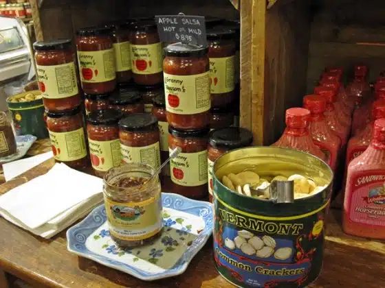 A display of preserves and condiments including apple salsa jars, horseradish bottles, an open jar with dip, and a can of common crackers on a shelf in a store.