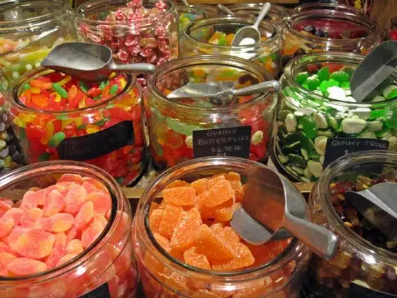 A variety of colorful candies in glass jars with scoops. Labels identify the assortment, including gummy butterflies, green gummy frogs, and orange gummies.