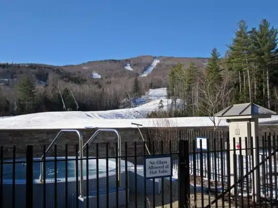 Outdoor hot tub enclosed by a black fence with a "No Glass Allowed at Hot Tubs" sign, overlooking snowy slopes and forested hills under a clear blue sky.