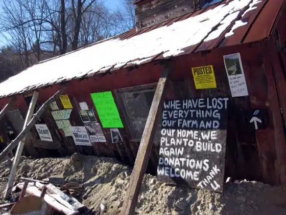 A damaged, leaning building with various signs attached. One sign reads, "We have lost everything, our farm, and our home. We plan to build again. Donations welcome." Snow is on the roof.