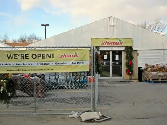 Exterior of a Shaw's supermarket with a sign on a fence reading "WE'RE OPEN!" and "Seafood, Fresh Produce, Fresh Dairy, Groceries And More!" The entrance is decorated with holiday wreaths.
