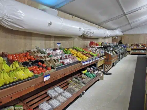 A grocery store aisle features various fruits and vegetables displayed in wooden bins under a white ceiling.