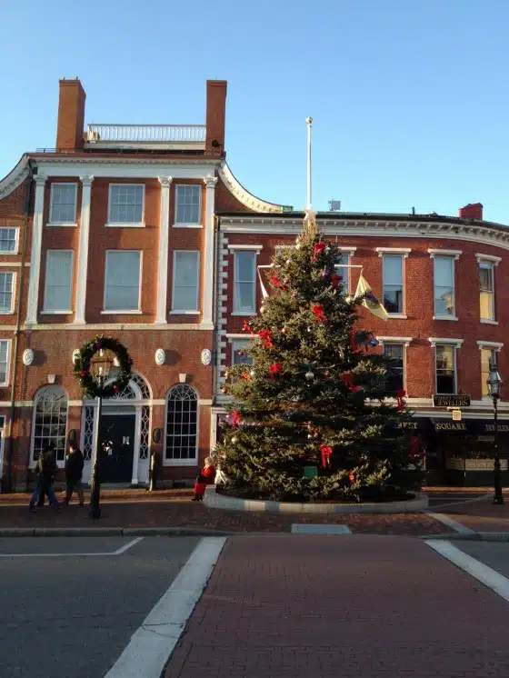 A decorated Christmas tree is placed in front of a historic brick building on a clear day. Pedestrians and a sidewalk are visible, along with a street in the foreground.