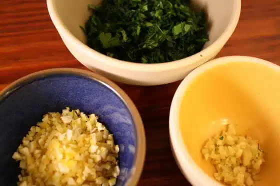 Chopped herbs and spices in three bowls on a wooden surface.