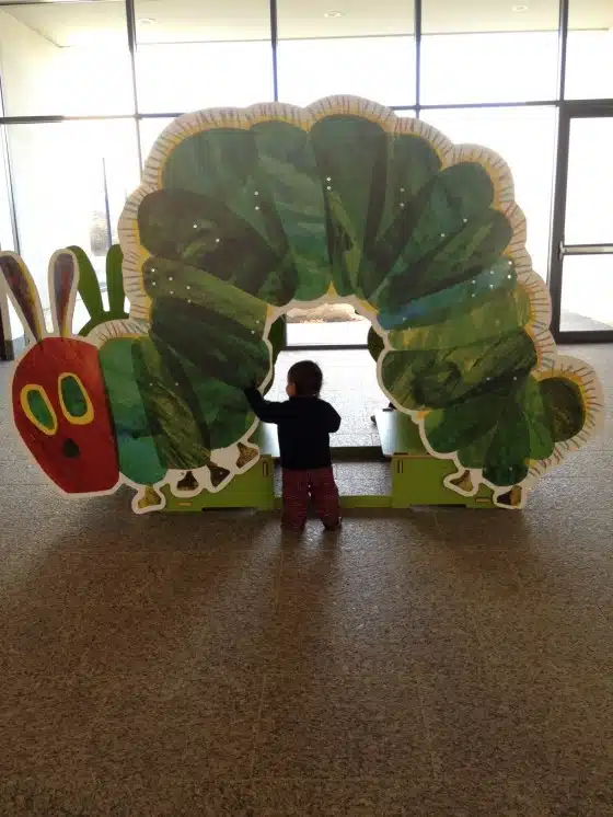 A child interacts with a large cutout of a green caterpillar in a brightly lit room with large windows.