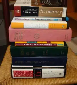 A stack of seven books on language and writing, including dictionaries, style guides, and a thesaurus, placed on a chair seat.