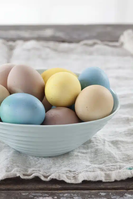 A light blue bowl filled with various colored eggs, including shades of blue, yellow, and brown, sits on a cloth-covered wooden surface.