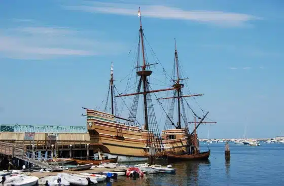 A wooden sailing ship docked at a pier in a harbor, with smaller boats nearby and a clear blue sky in the background.